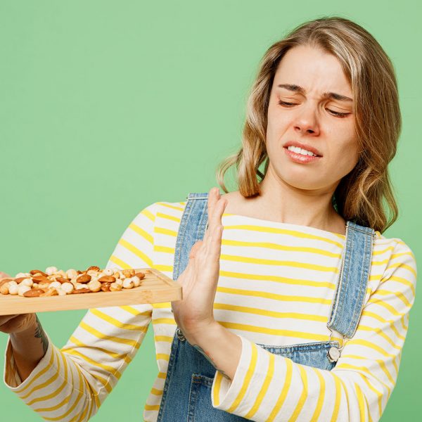 Sick ill allergic woman has red watery eyes runny stuffy sore nose suffer from allergy trigger symptoms hay fever hold peanuts nuts show stop gesture isolated on plain green background studio portrait